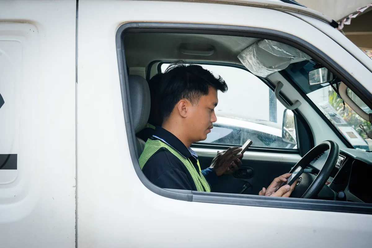 A technician at the wheel of a parked work van checking a phone, a colleague in the passenger seat doing the same.