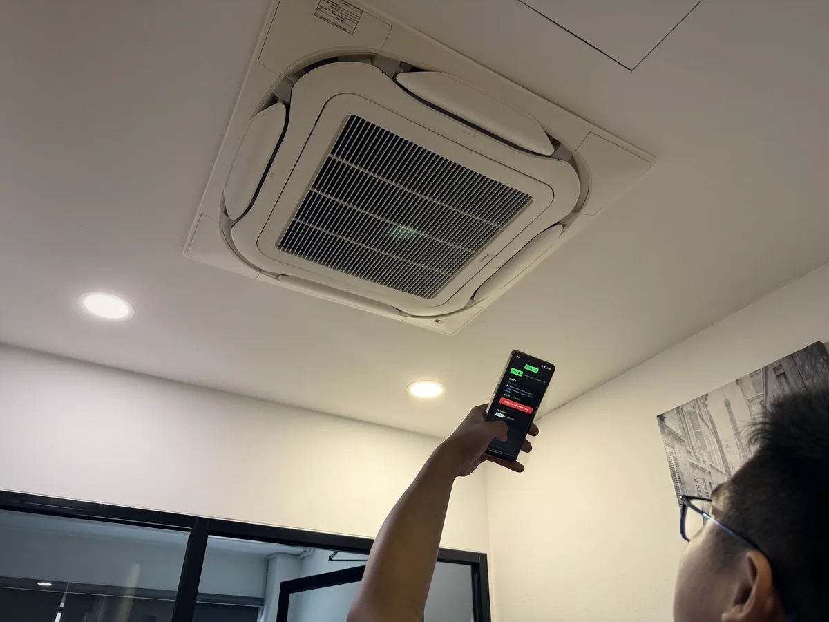 A technician under a ceiling cassette unit, holding up a phone with the Technician Portal's visit steps on screen.